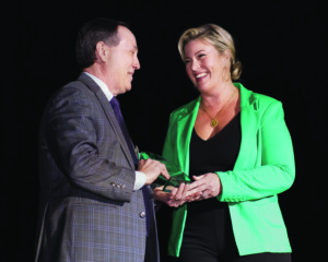 A woman with blonde hair in a green blazer presents a white man in a dark suit an award.